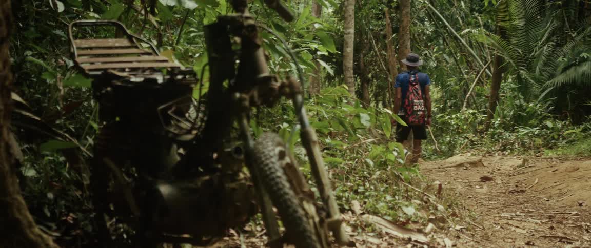A hiker with a backpack walks down a dirt trail in the jungle, passing an old rusted motorcycle abandoned in the trees