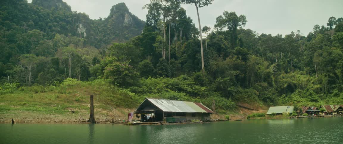 A rustic floating house sits peacefully on a tranquil lake at the edge of a lush, dense jungle