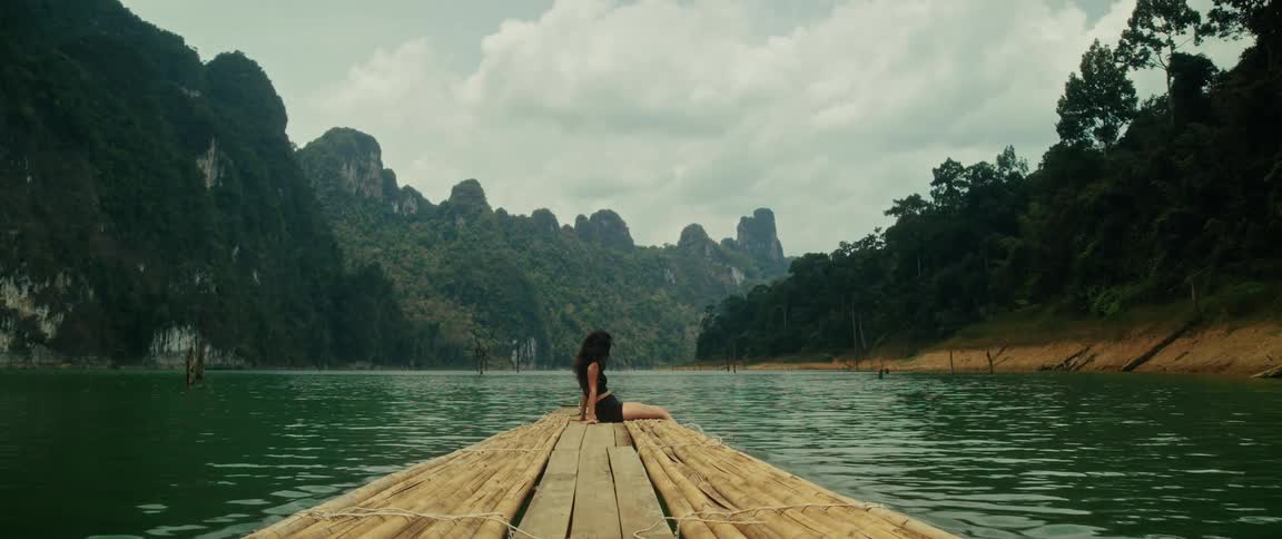 A woman sits peacefully on the edge of a bamboo raft as it floats on a serene lake surrounded by lush green mountains