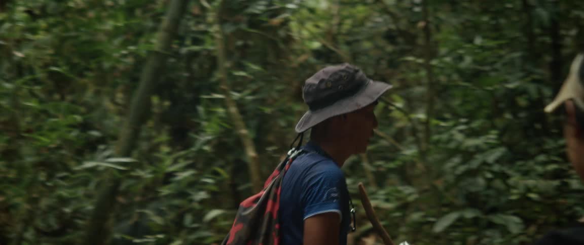 A jungle guide with a walking stick leads a hiker up a steep and muddy trail in slow motion