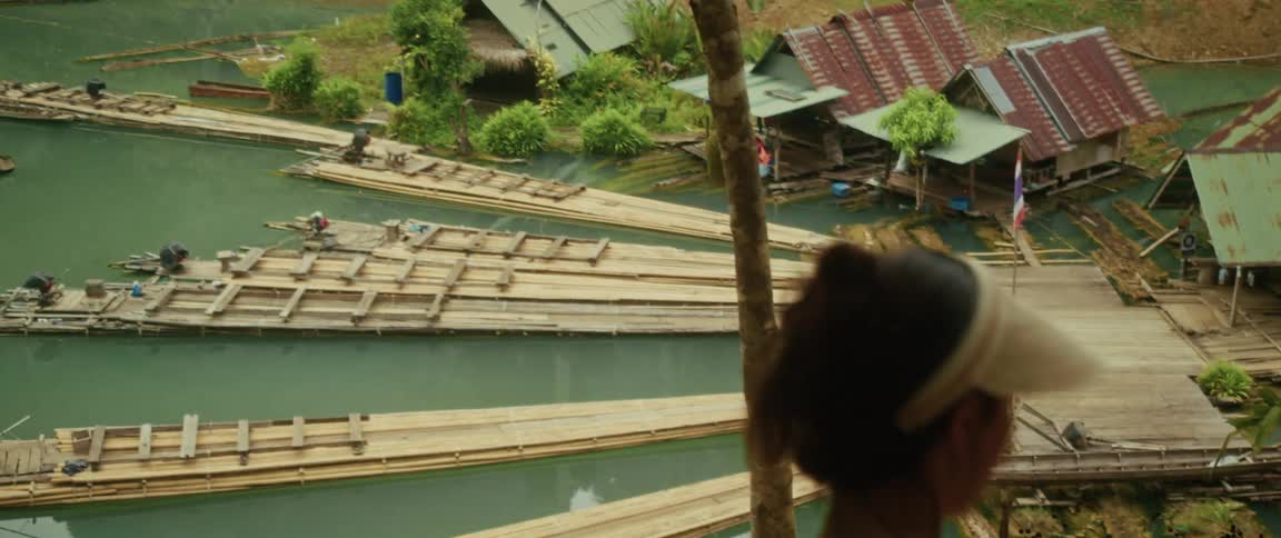 A traveler looks down from a viewpoint at a bustling riverside village with bamboo rafts in Thailand
