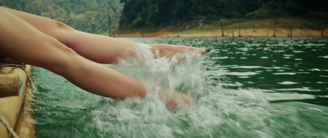 A person's bare feet splash in a green jungle lake from the side of a moving boat in slow motion