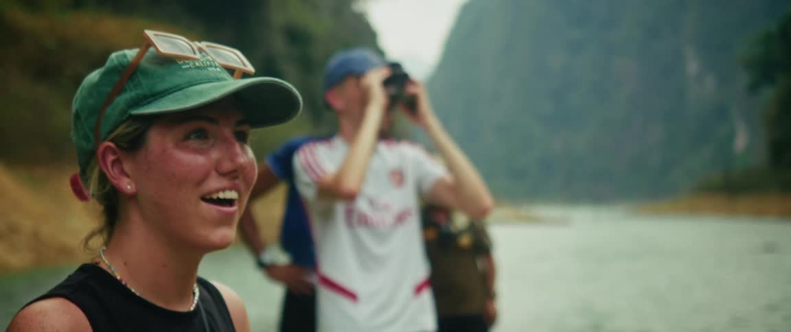 A joyful young woman points out a discovery while on a boat in a beautiful mountain lake, as her friend looks on with binoculars