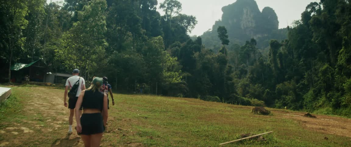 A group of friends in slow motion walks along a path into a lush tropical jungle towards a towering mountain