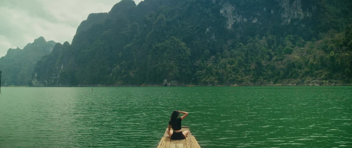 A woman on a bamboo raft peacefully enjoys the breathtaking scenery of a tranquil, emerald-green lake and lush tropical mountains