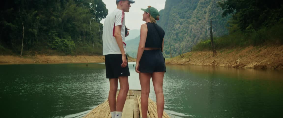 A young couple holds hands while enjoying a romantic journey on a bamboo raft down a tranquil lake surrounded by lush green mountains