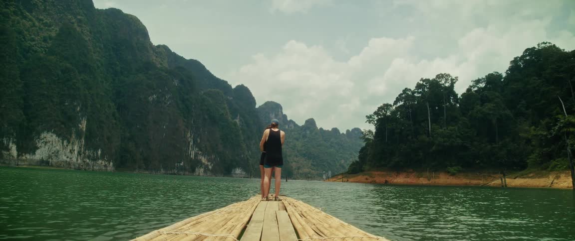 A loving couple embraces on the front of a bamboo raft while floating through a tranquil lake surrounded by majestic green mountains