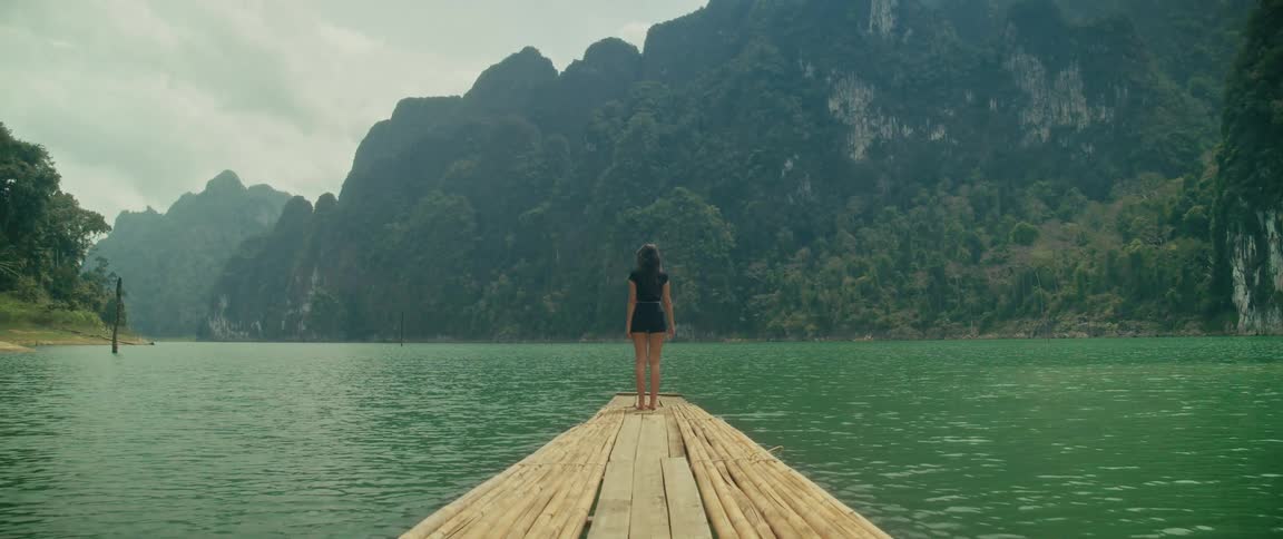 A woman walks barefoot to the end of a bamboo raft on a tranquil lake, surrounded by majestic green mountains