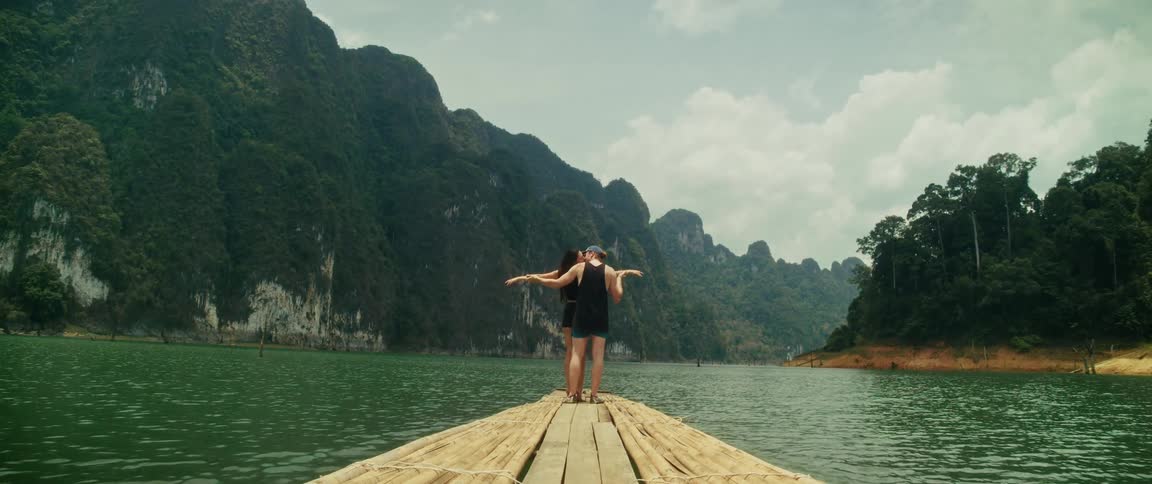A romantic couple embraces and kisses while standing at the front of a bamboo raft on a serene lake surrounded by lush green mountains