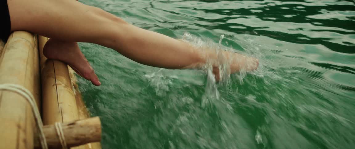 Close-up slow motion of a woman's feet splashing playfully in the green lake water from a bamboo raft