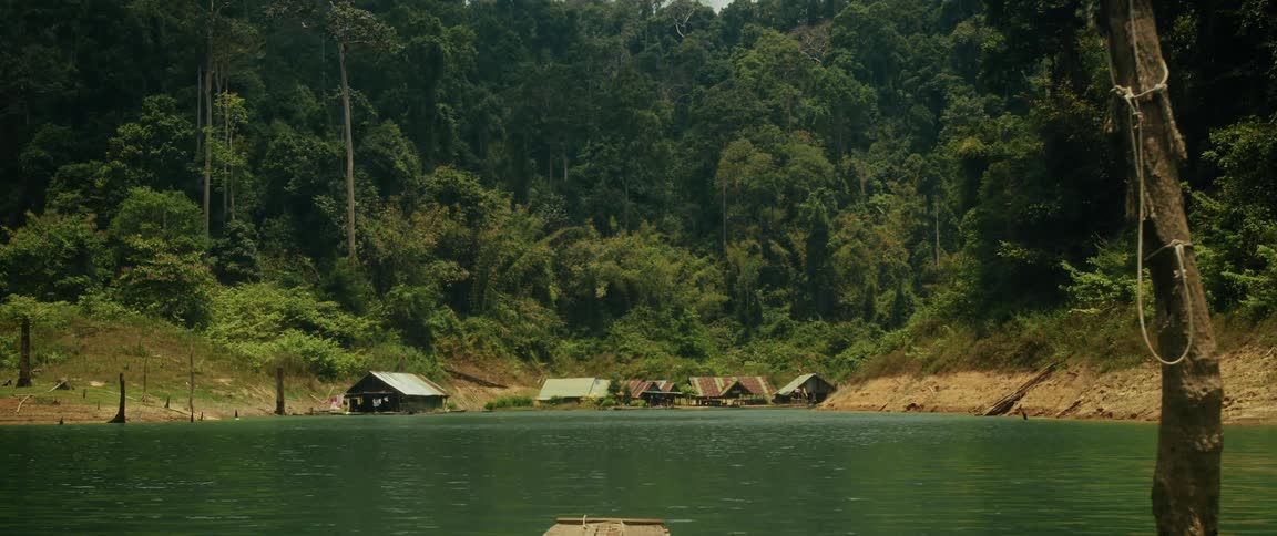 A serene view from a wooden raft of a remote village with floating huts on a tranquil lake surrounded by a dense jungle