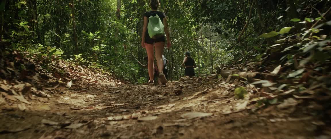 A group of friends hike up a steep, leafy trail in a dense tropical jungle in slow motion