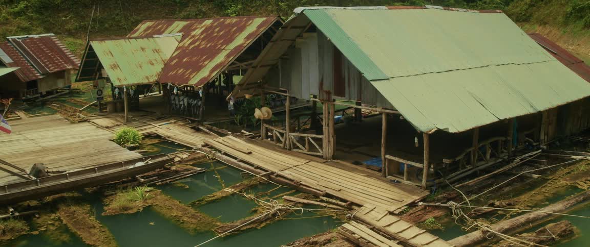 Rustic wooden floating houses with weathered tin roofs sit peacefully on a serene green river in a lush jungle