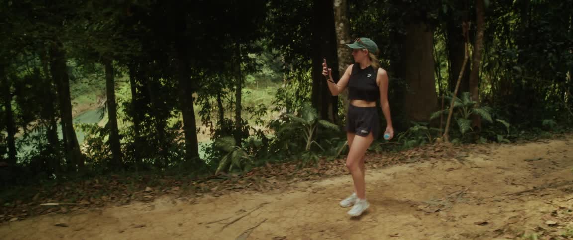 A young woman in athletic wear walks along a jungle trail, filming the lush scenery with her smartphone