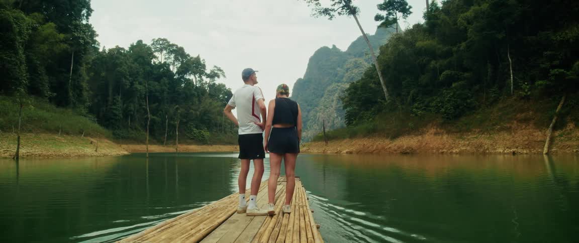 A young couple stands on the front of a bamboo raft, holding hands and admiring the scenic views as they float down a tranquil river through a lush tropical jungle