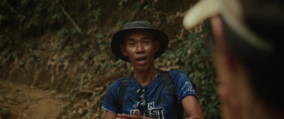 A friendly Asian tour guide with a hat and backpack enthusiastically explains something to a tourist in a lush jungle setting