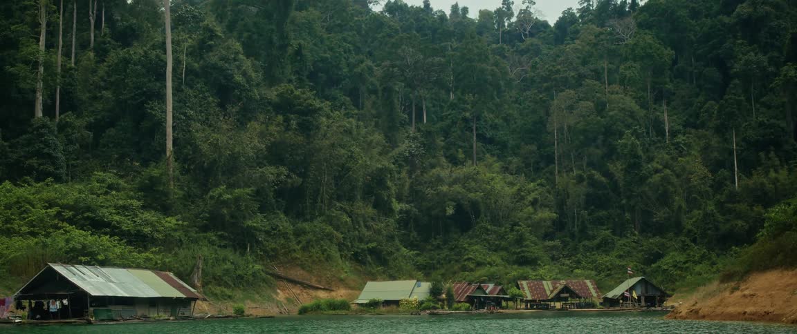 A tranquil floating village of rustic bungalows rests on a calm lake at the edge of a dense, vibrant green jungle