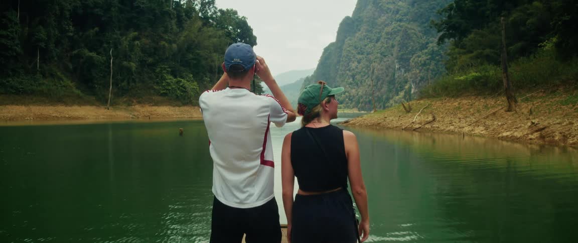 A young couple on a bamboo raft floats down a tranquil green river, pointing and using binoculars to explore the stunning tropical jungle and mountains around them