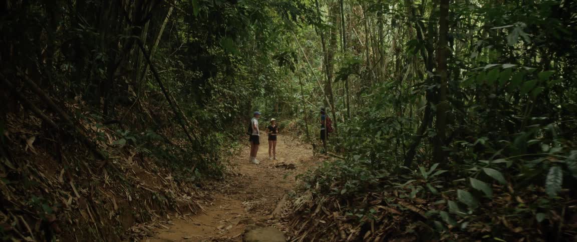 A tilt-down shot reveals three young hikers taking a break on a dirt trail deep within a lush, green tropical jungle
