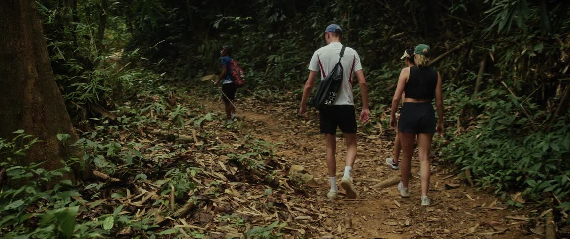 A group of friends follow a local guide as they hike up a dirt trail through a lush, dense jungle
