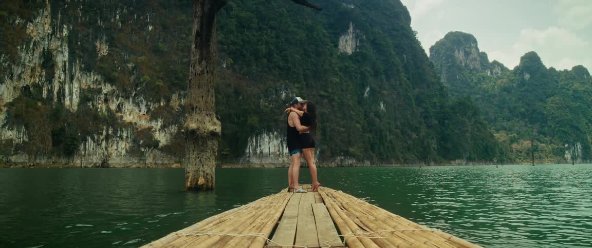 A loving couple shares a romantic kiss on the bow of a bamboo raft as it floats across a tranquil lake surrounded by majestic mountains