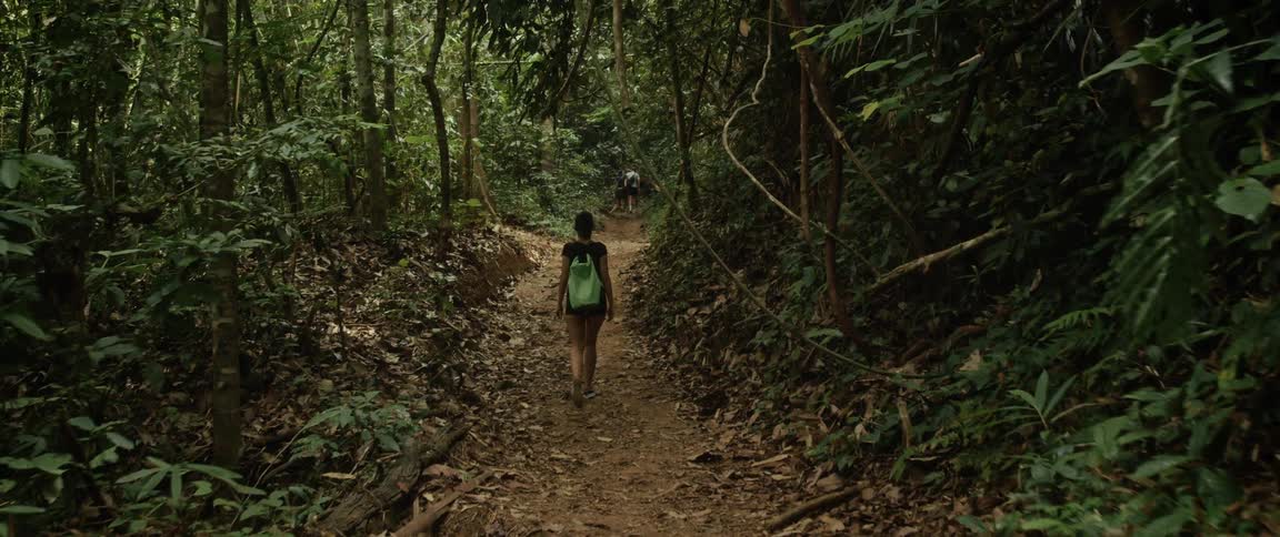 A woman with a backpack hikes along a dirt trail through a dense tropical rainforest in slow motion