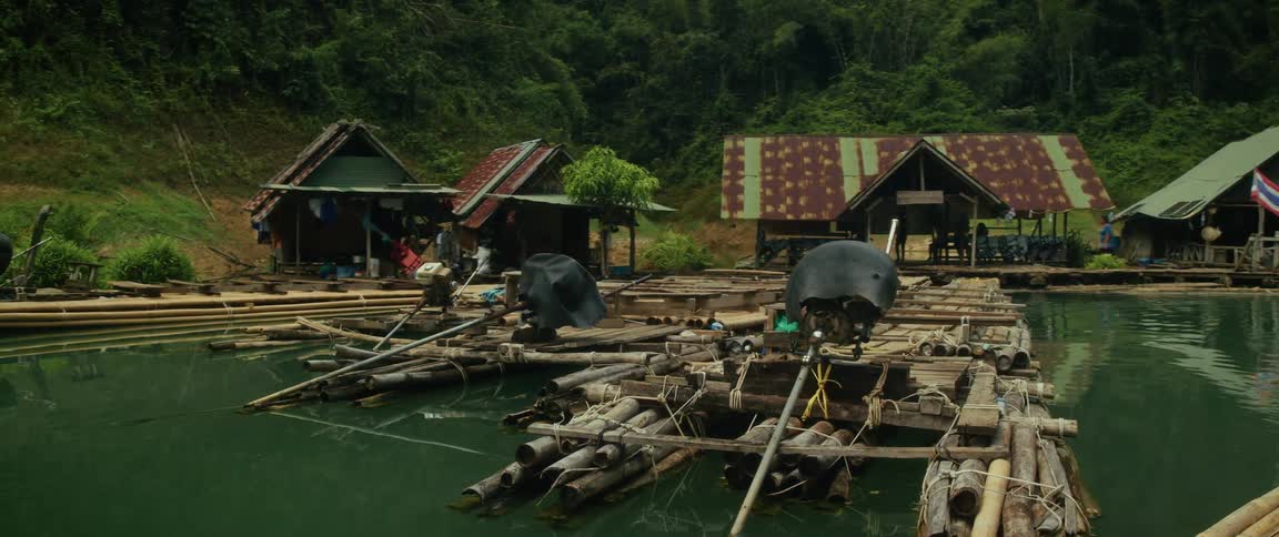 Slow pull-back shot reveals a rustic floating village with bamboo rafts and huts nestled by a lush jungle hillside on a calm lake in Thailand