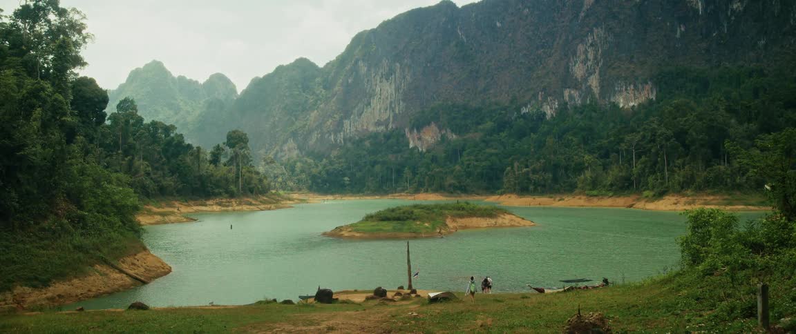 A beautiful wide shot of a tranquil lake surrounded by lush green mountains, with a small island in the center and people on the shore