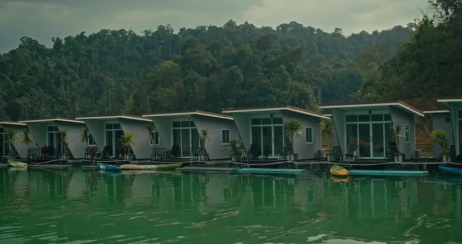 A line of modern floating bungalows with colorful kayaks sits on a calm green lake at the edge of a lush tropical forest