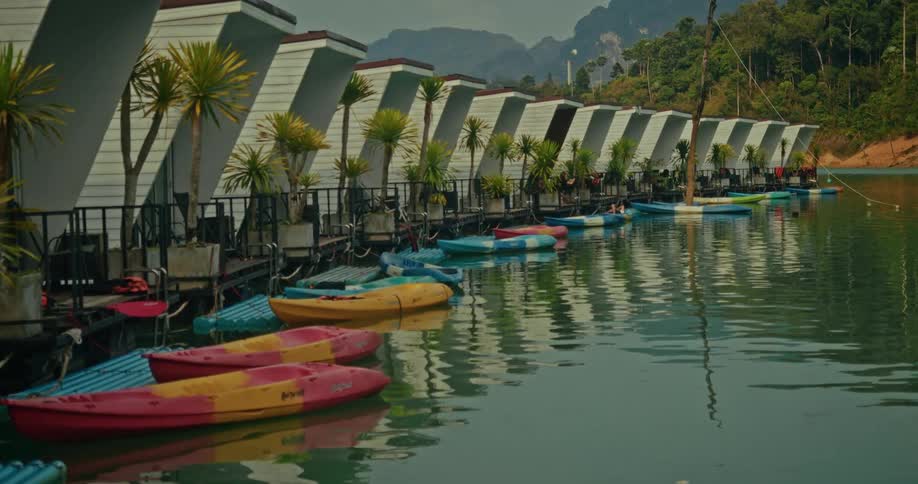 A tranquil lakeside resort features a row of modern floating cabins with colorful kayaks docked in the calm water