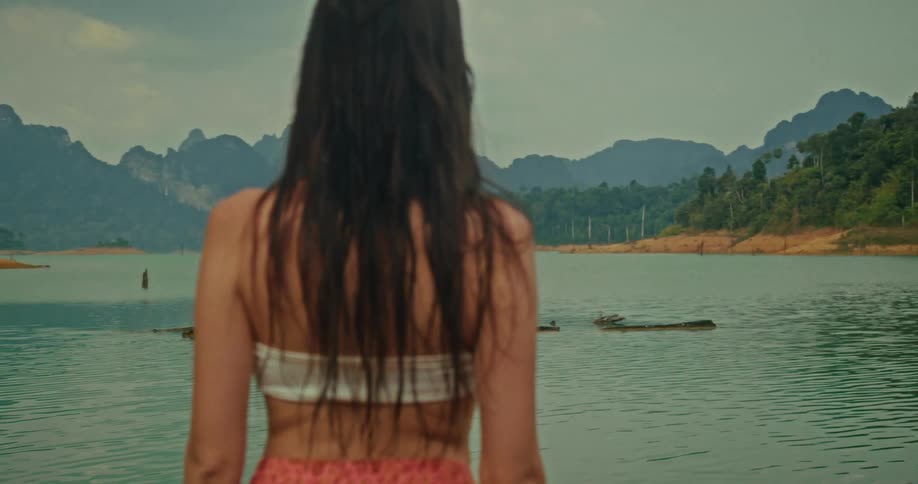 The camera slowly pushes past a woman standing at the edge of a serene mountain lake, revealing the vast, beautiful landscape