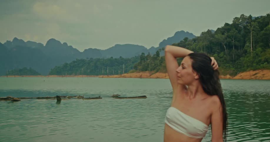 A beautiful woman sits by a tranquil mountain lake, sensually running her hand through her long, wet hair in slow motion