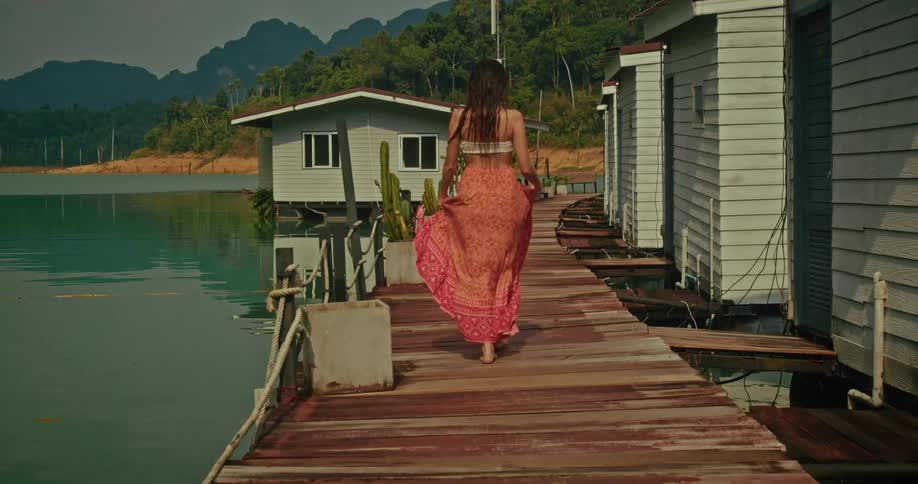 A woman in a beautiful flowing dress walks barefoot and dances on a wooden pier next to floating bungalows on a tranquil tropical lake