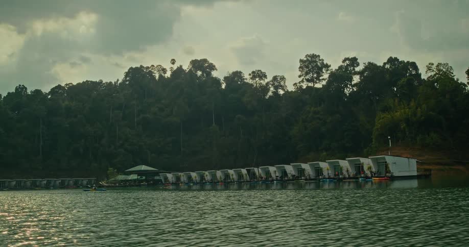 A wide, tranquil shot of a modern floating resort with bungalows lining a calm lake against a lush, jungle-covered hillside