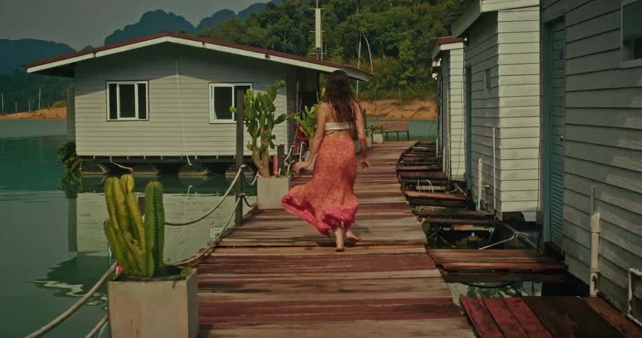 A carefree woman in a long flowing skirt runs barefoot down a wooden pier alongside floating bungalows on a tropical lake