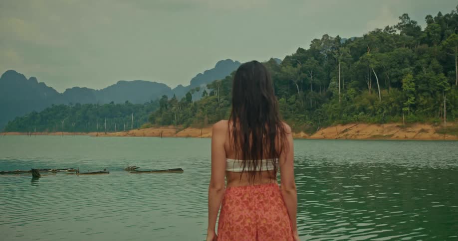 A pensive woman with long dark hair stands on a dock looking out at a serene lake surrounded by lush mountains