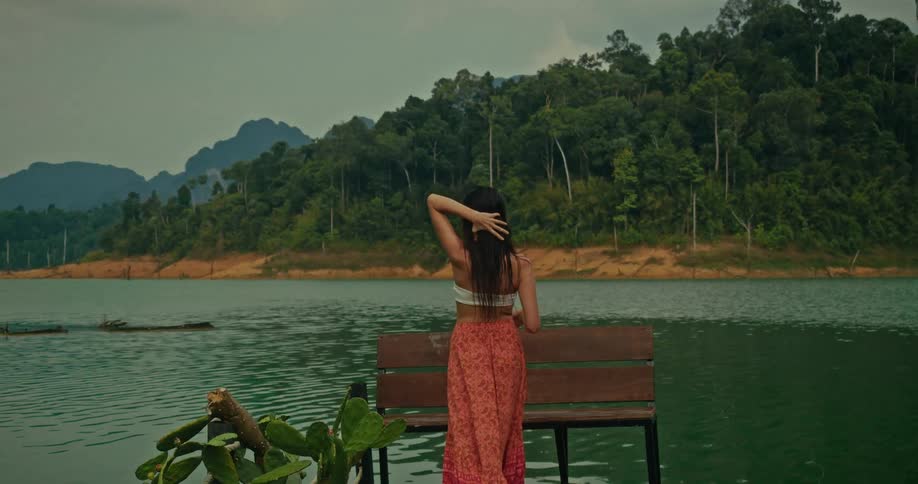 A smiling woman with long dark hair walks in slow motion along a pier, taking in the serene view of a vast lake and lush green mountains
