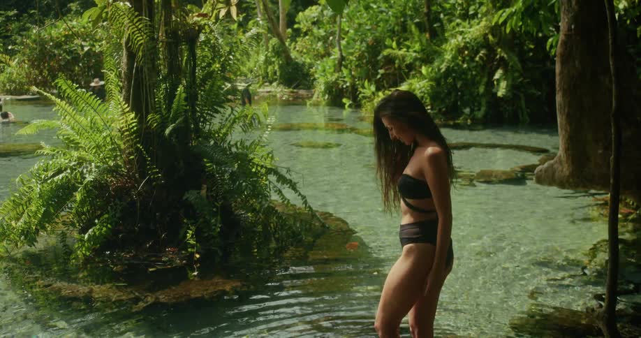 A beautiful woman in a black bikini stands in the shallow, clear water of a tranquil jungle spring