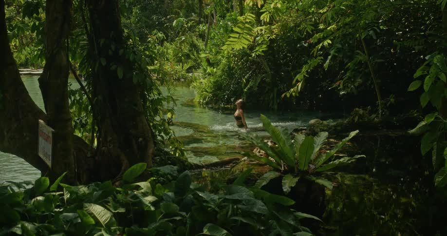 A woman in a black bikini flips her long wet hair back in slow motion while swimming in a pristine jungle spring
