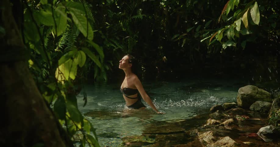 A serene woman in a bikini enjoys a refreshing dip in a clear jungle stream, flipping her hair back in the sunlight
