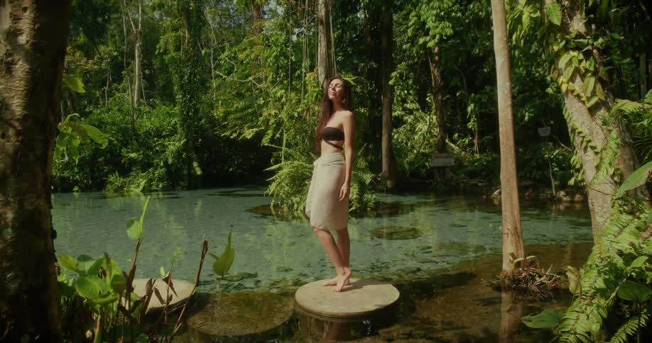 A serene woman in a sarong finds tranquility while standing on a stepping stone in a vibrant blue jungle pond surrounded by lush greenery