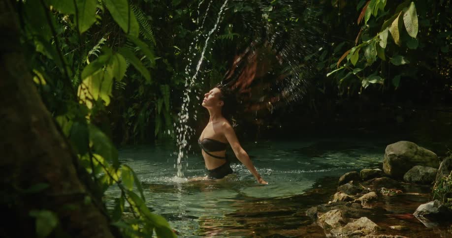 A beautiful woman in a black bikini dramatically flips her long wet hair back in slow motion while bathing in a secluded jungle spring