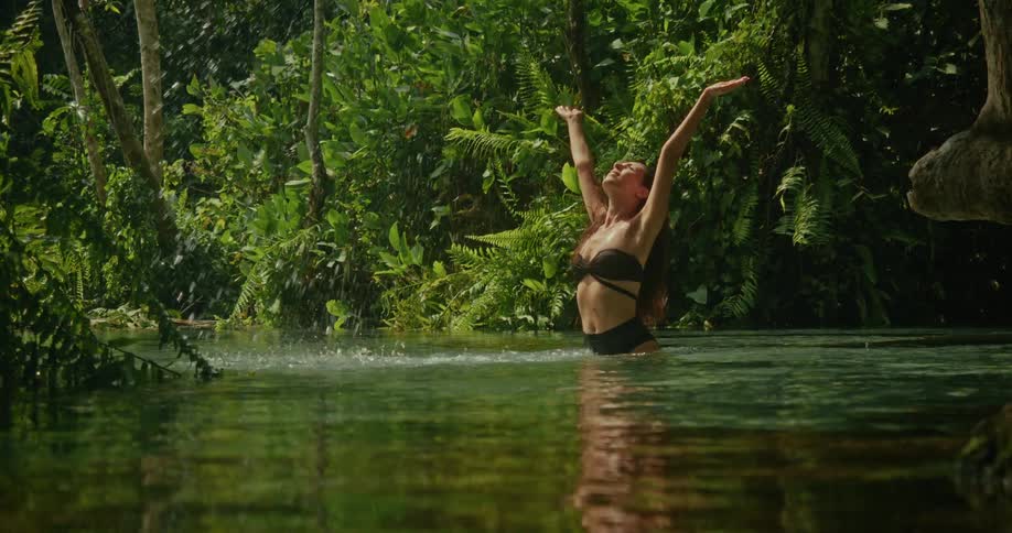 A happy woman in a bikini splashes water and raises her arms to the sky while bathing in a tranquil jungle spring