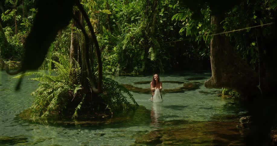 A happy woman in a bikini playfully splashes water in a crystal-clear jungle spring, surrounded by lush tropical foliage