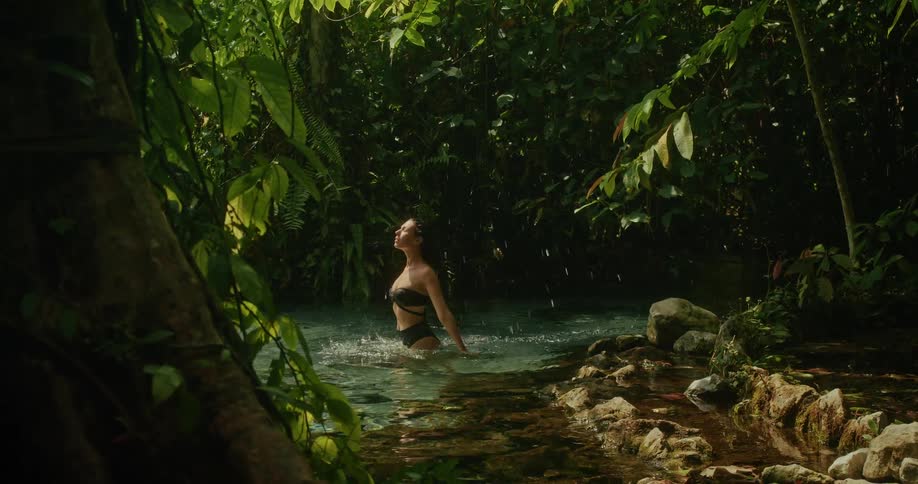 A beautiful woman in a black bikini flips her wet hair back in slow motion while bathing in a pristine, sun-dappled jungle spring