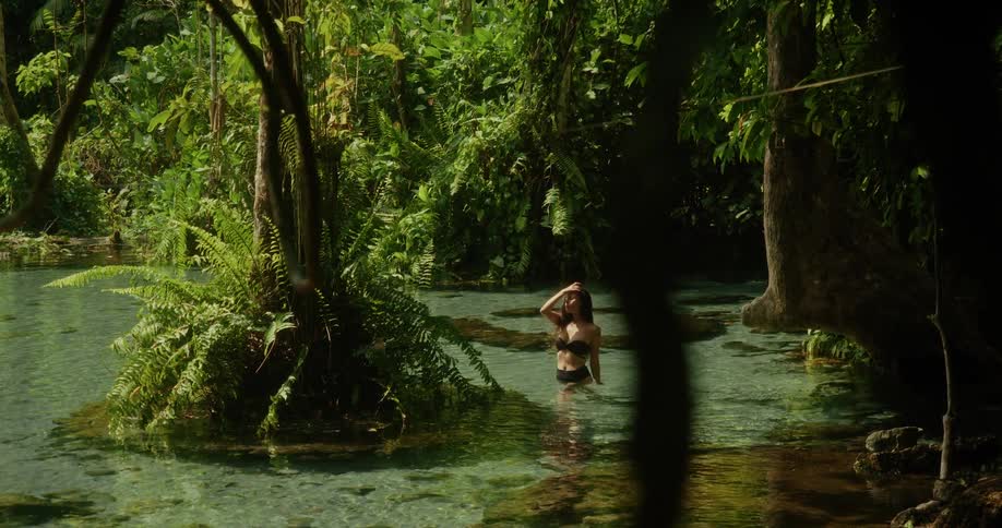 A beautiful woman in a black bikini soaks in the sun while wading through a crystal clear spring in a lush, tropical jungle