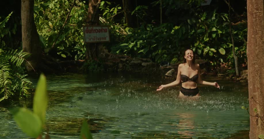 A smiling woman in a bikini playfully splashes water in a beautiful, sunlit jungle spring in slow motion