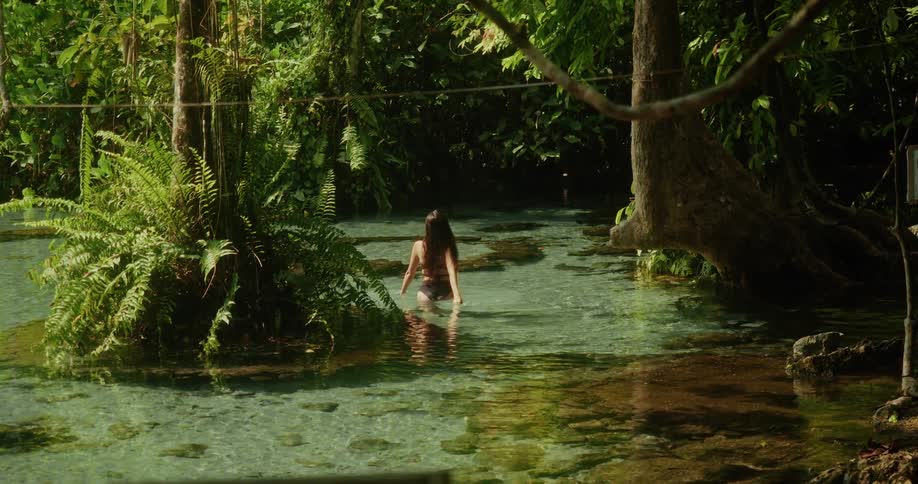 A young woman in a black bikini wades through the shallow, crystal-clear waters of a secluded jungle oasis, surrounded by lush greenery
