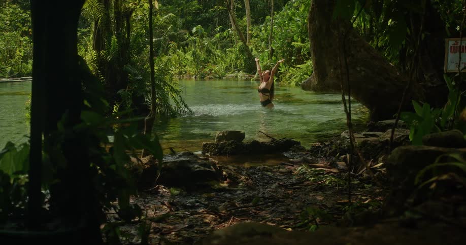 A joyful woman in a bikini splashes and raises her arms in the clear turquoise water of a secluded jungle river