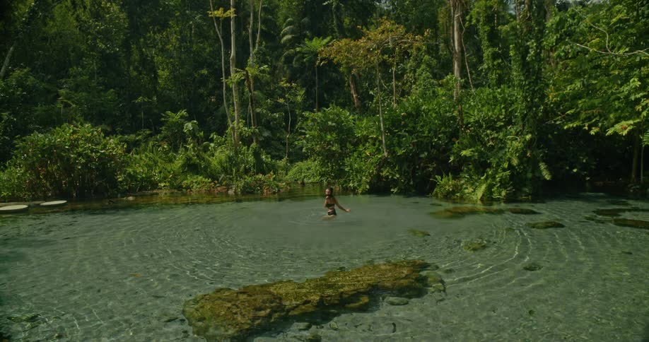 A woman wades into the crystal-clear waters of a serene jungle spring, surrounded by lush tropical rainforest